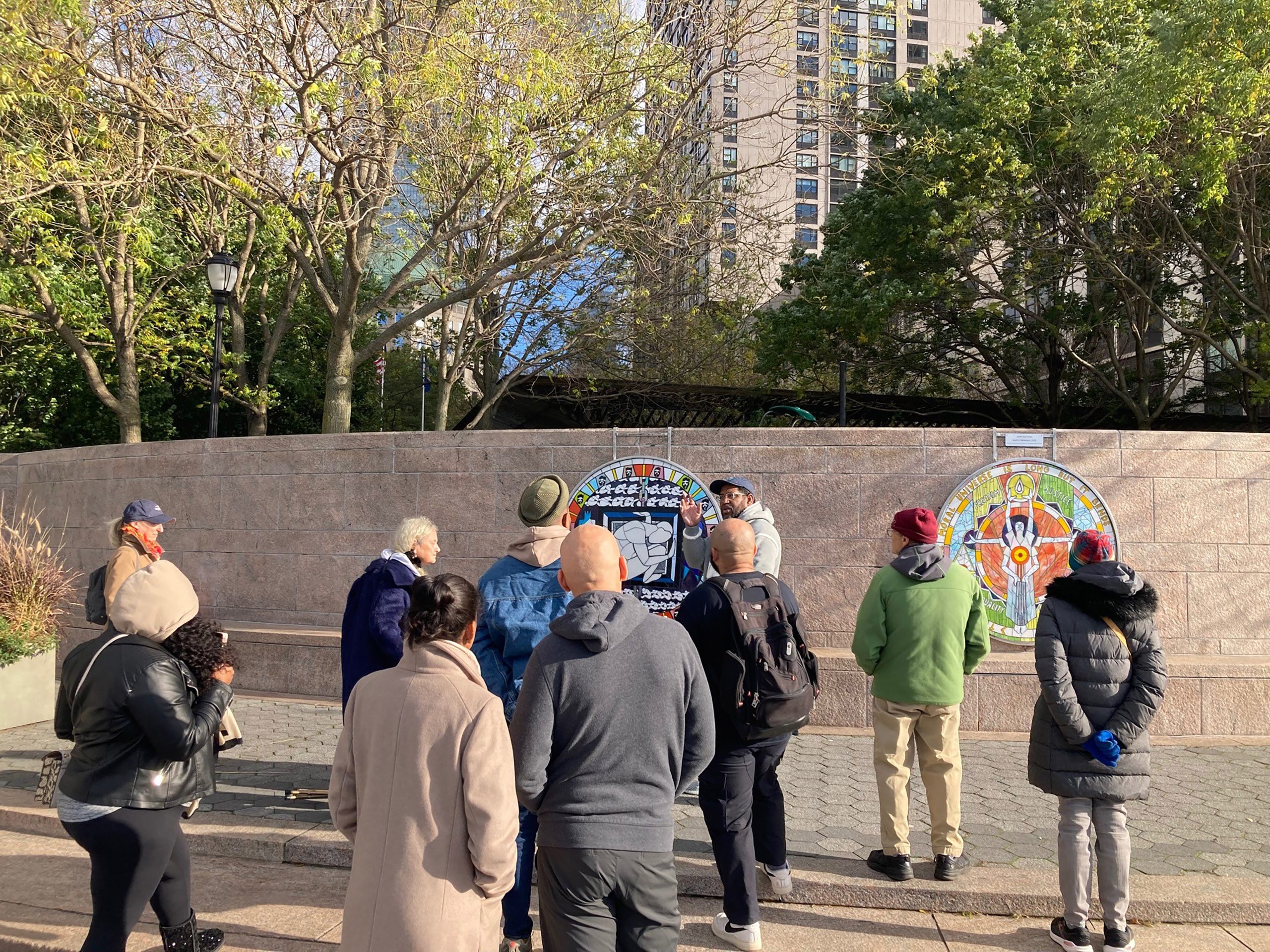 A photograph of the artist James Yaya Hough, a Black man in glasses and a blue cap, speaking to a crowd of people in a park in front of his public artwork Justice Reflected.