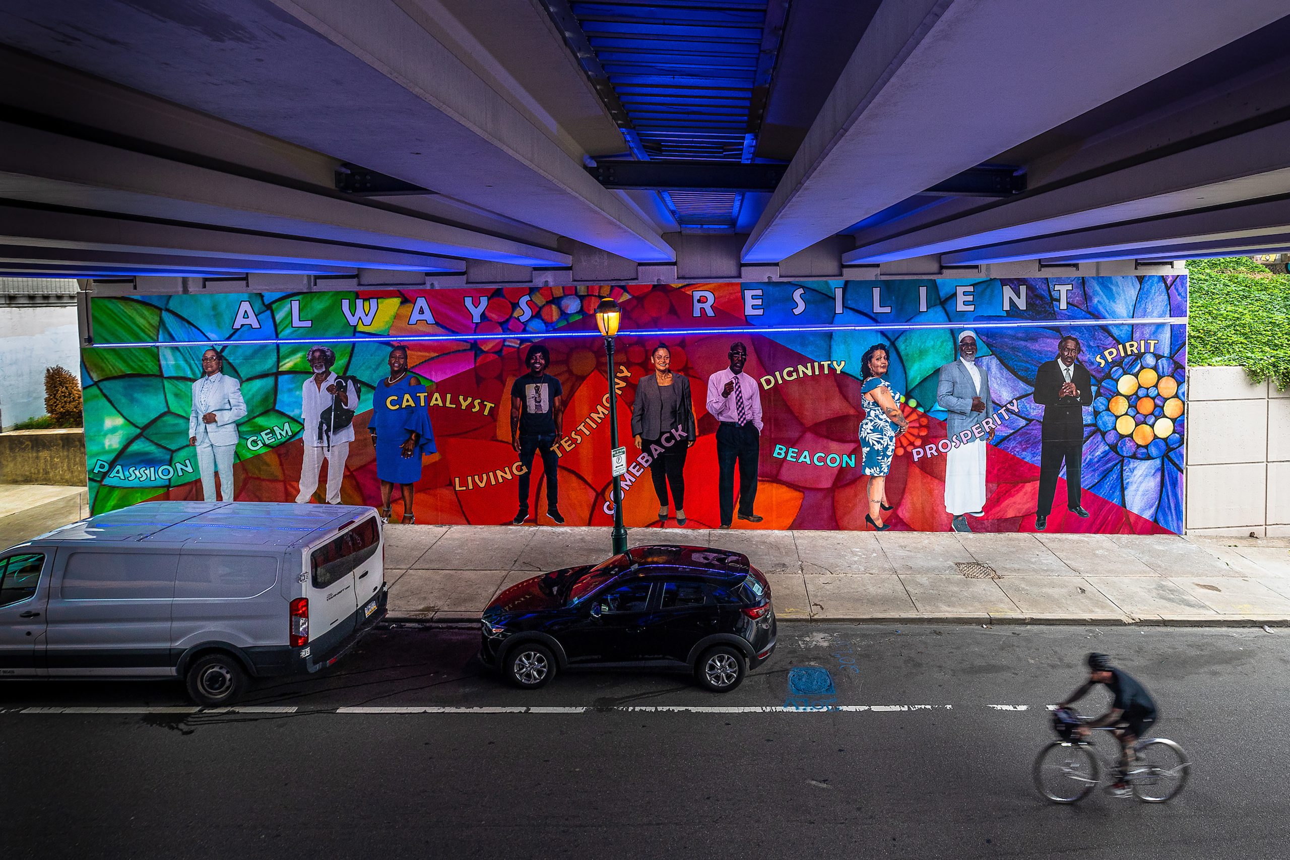 A public mural facing a street shows Black men and women standing in bright clothing against a background of a brightly colored floral pattern with words capturing resilience integrated into the pattern. Above them are the words 