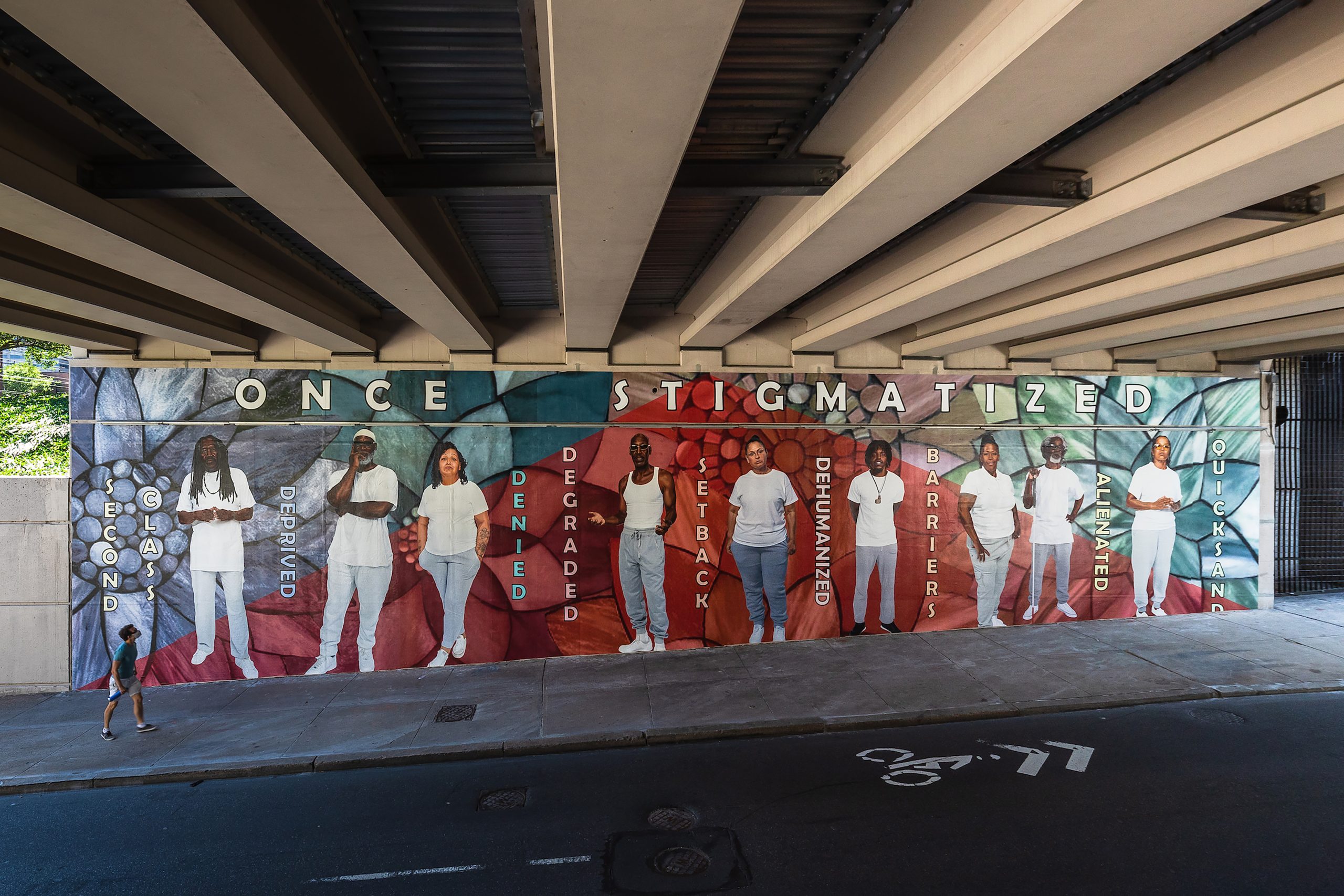 A public mural facing a street shows Black men and women standing in pale clothes against a background of floral shapes in gray, with a triangle of transparent red layered over it, and words capturing stigma. Above them are the words 