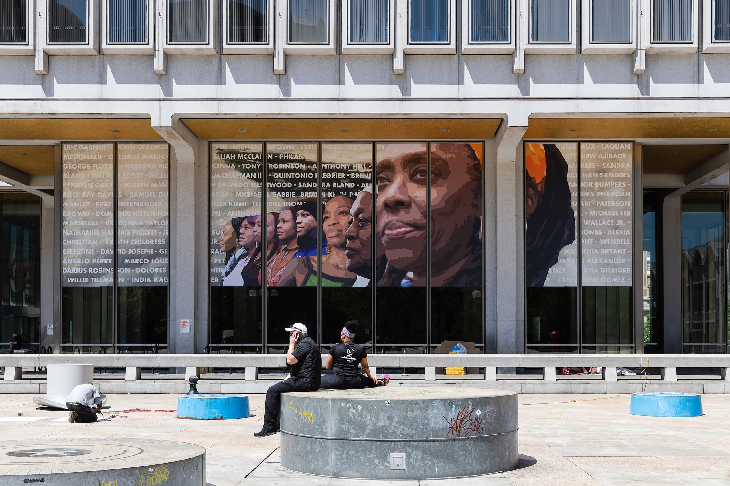 A public mural depicts Black women activists standing in a line of strength appear against a background showing the names of people recently killed by police brutality.