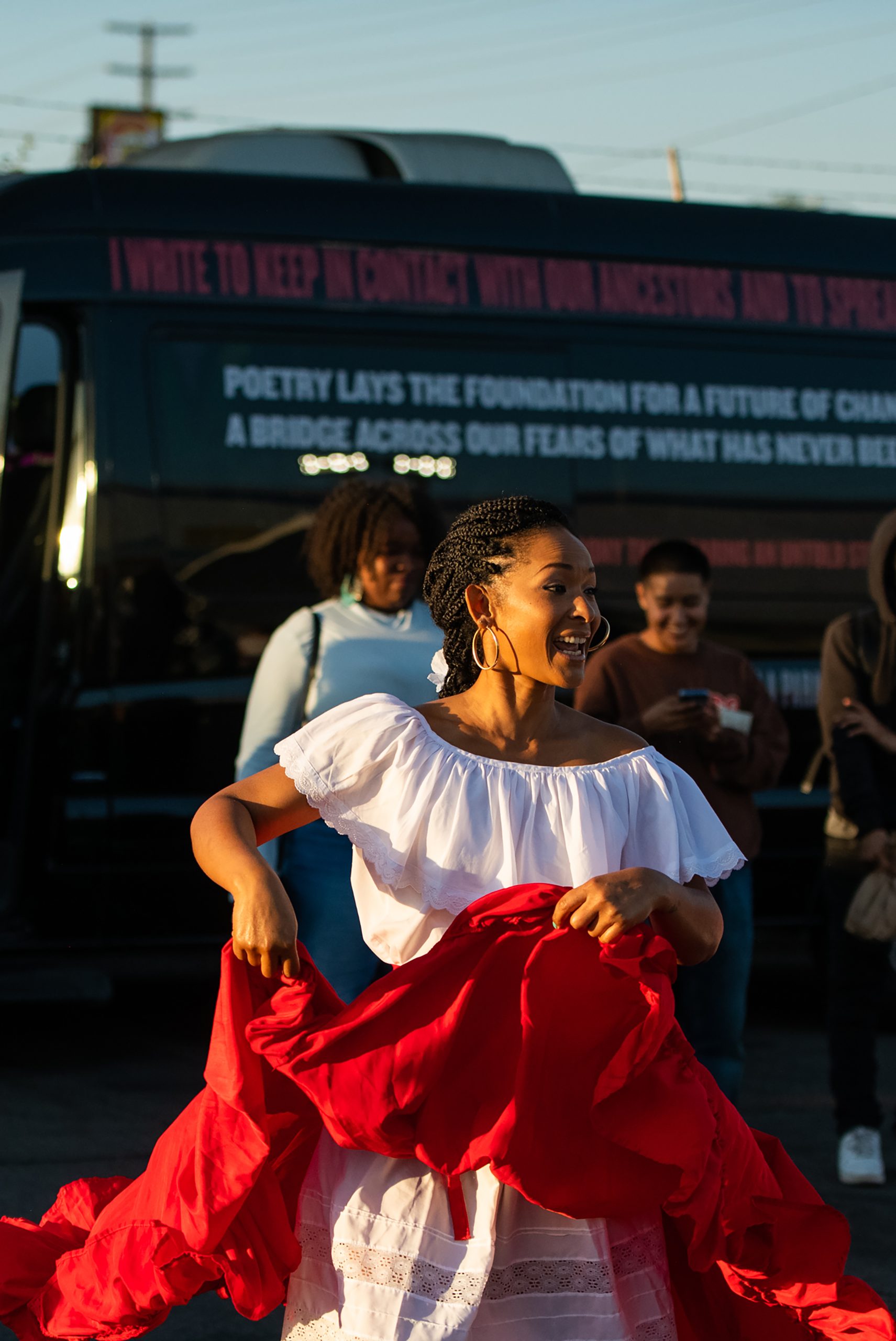 A photograph of Nadia Calmet, a woman of Afro-Peruvian descent wearing a white top and bright red overskirt that she holds up, smiles as she performs a dance for an audience outside.